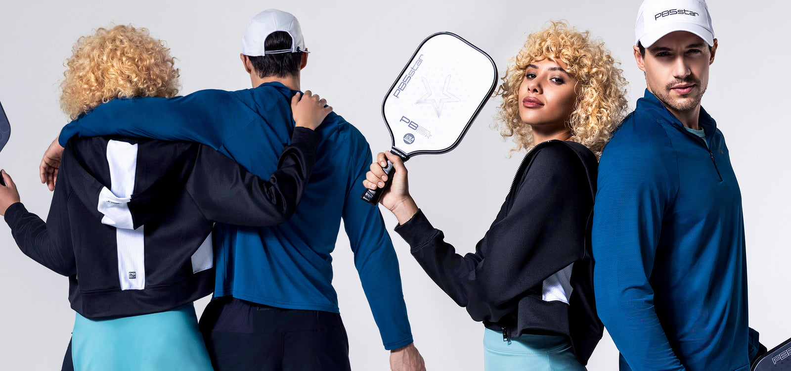 Two models wearing PB5 activewear, a woman wearing a black hoodie with white mesh detail and an arctic side split skirt, and a man with a blue long-sleeve quarter-zip top and white PB5star cap. Both hold PB5star pickleball paddles, showing a sporty and stylish look.