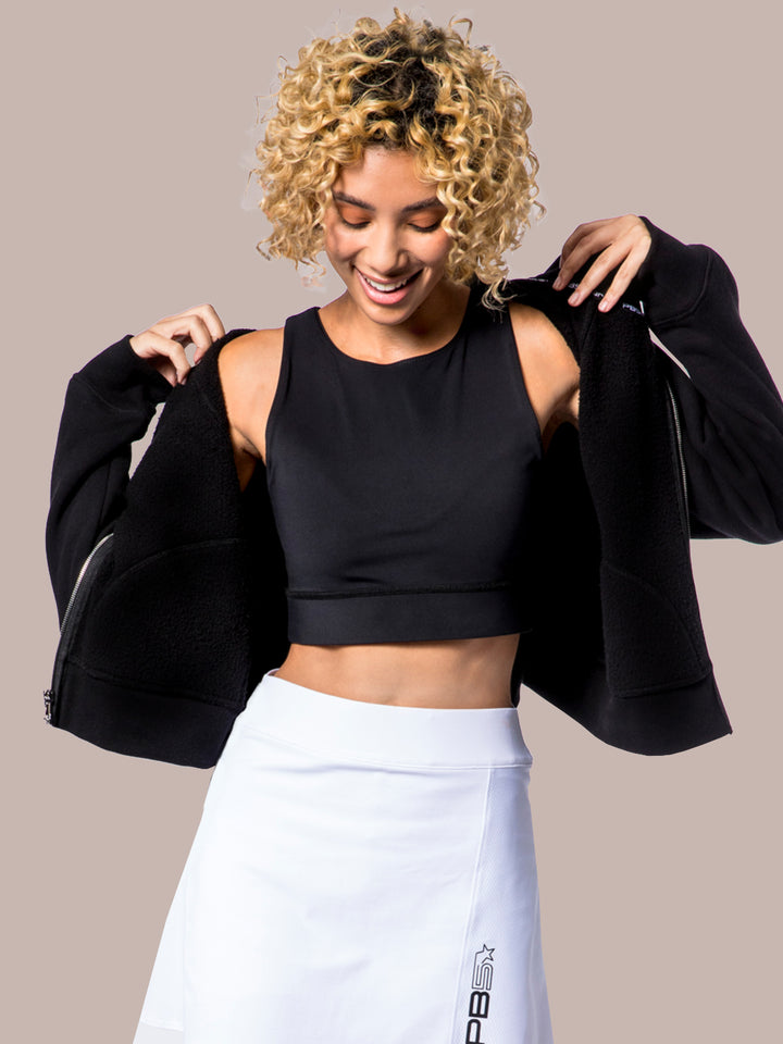 Joyful model putting on a PB5Star black Luxe Cropped Lounge Hoodie with a Mesh Panel Pickleball skirt for a modern athletic look.