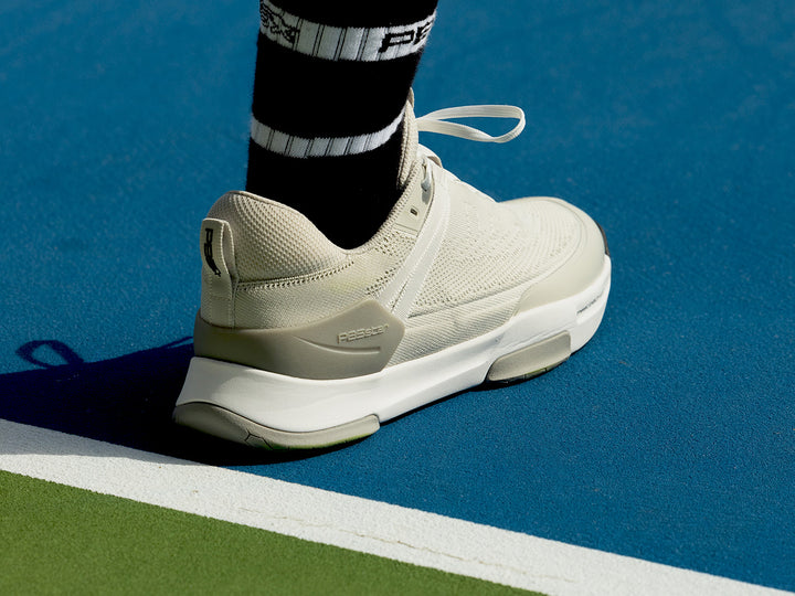 Close-up of the PB5 Court2 tennis shoe in light grey, showcasing its dynamic stability assist technology and sleek heel design, worn on a pickleball court with vibrant blue and green colors.