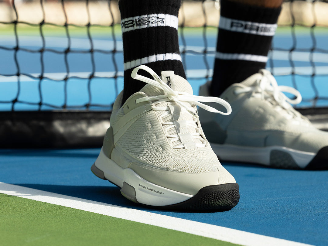 Close-up of PB5 Court2 performance tennis shoes in white, worn on a blue and green tennis court with a net in the background. Features dynamic stability assist technology for optimal court performance.