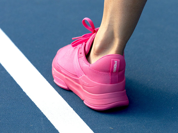 Rear view of a person wearing PB5 Court2 pink athletic shoes with PB5star branding, standing on a blue pickleball court.