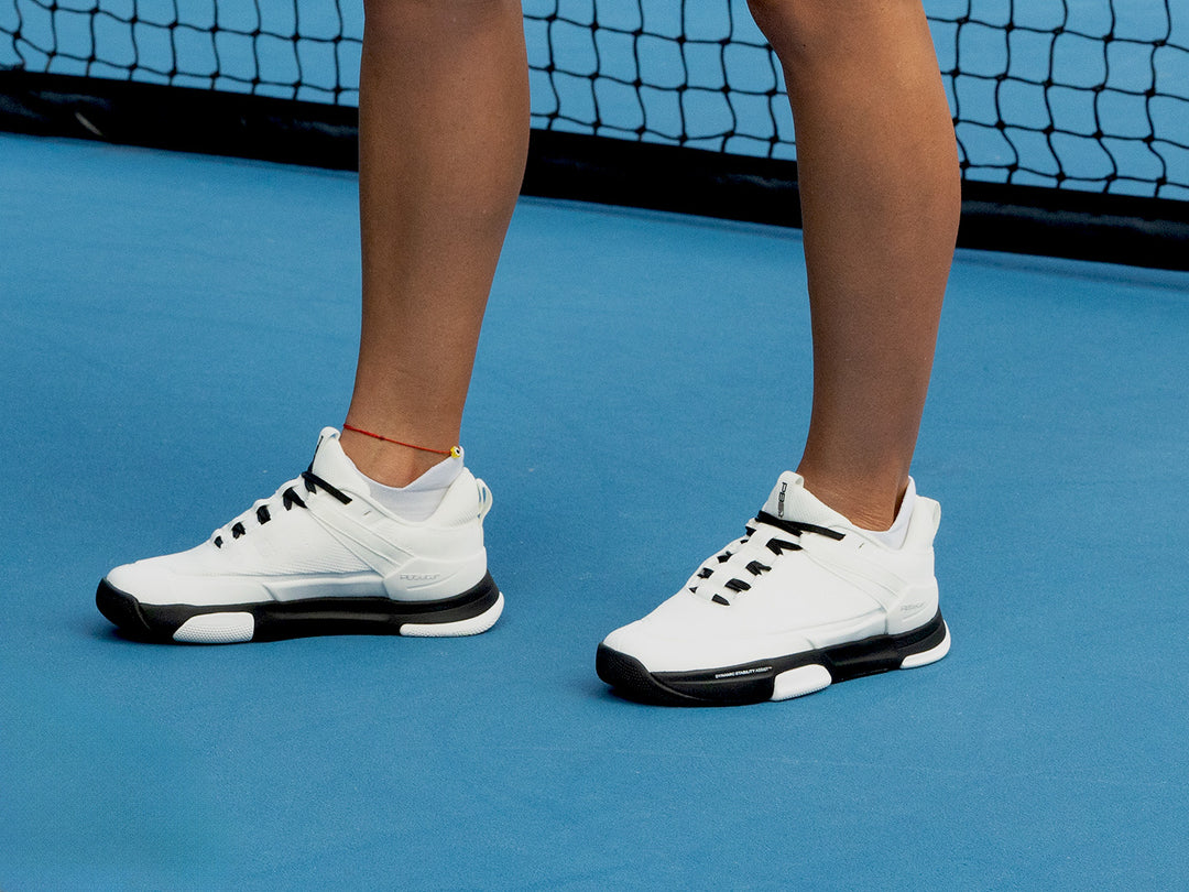 Person wearing PB5 Court2 white and black athletic shoes with PB5star branding, black laces, and black cushioned soles on a blue pickleball court.