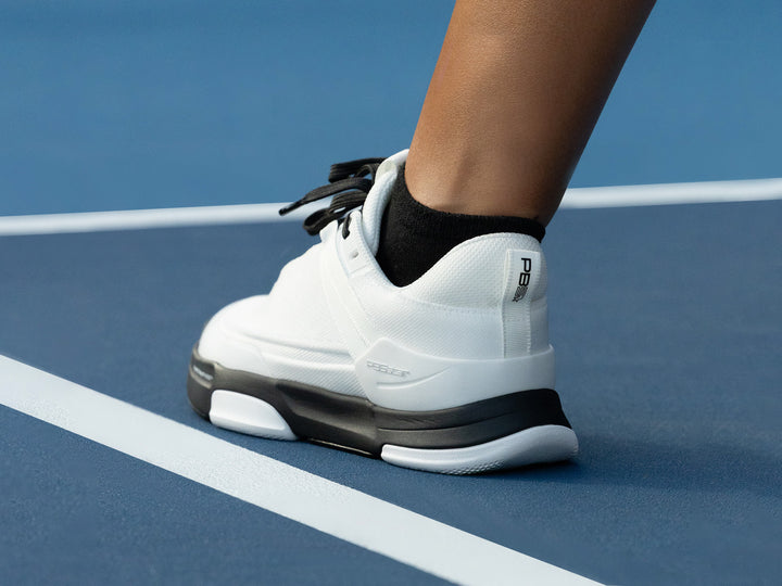 Player wearing PB5 Court2 athletic shoes in White and black with PB5star branding, stepping on a blue pickleball court.