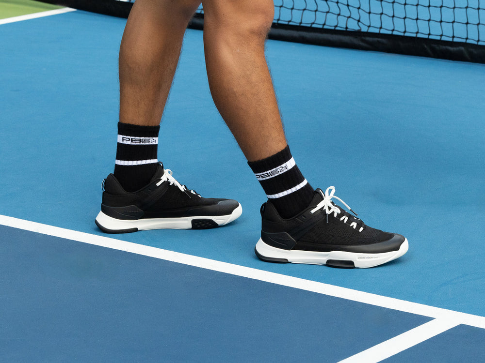 Male player wearing PB5 Court2 black athletic shoes with PB5star branding and black PB5star performance crew socks, walking on a blue pickleball court.